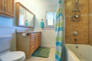 Sweet bathroom interior in soft blue and green tones. View of honey color cabinet with mirror shower and toilet.